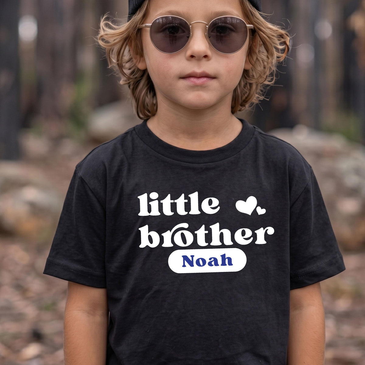 May include: A black short-sleeved t-shirt featuring the white text "little brother" with two small white hearts above it. Below the text is a white horizontal pill-shaped graphic containing the blue name "Noah" in a serif font.
