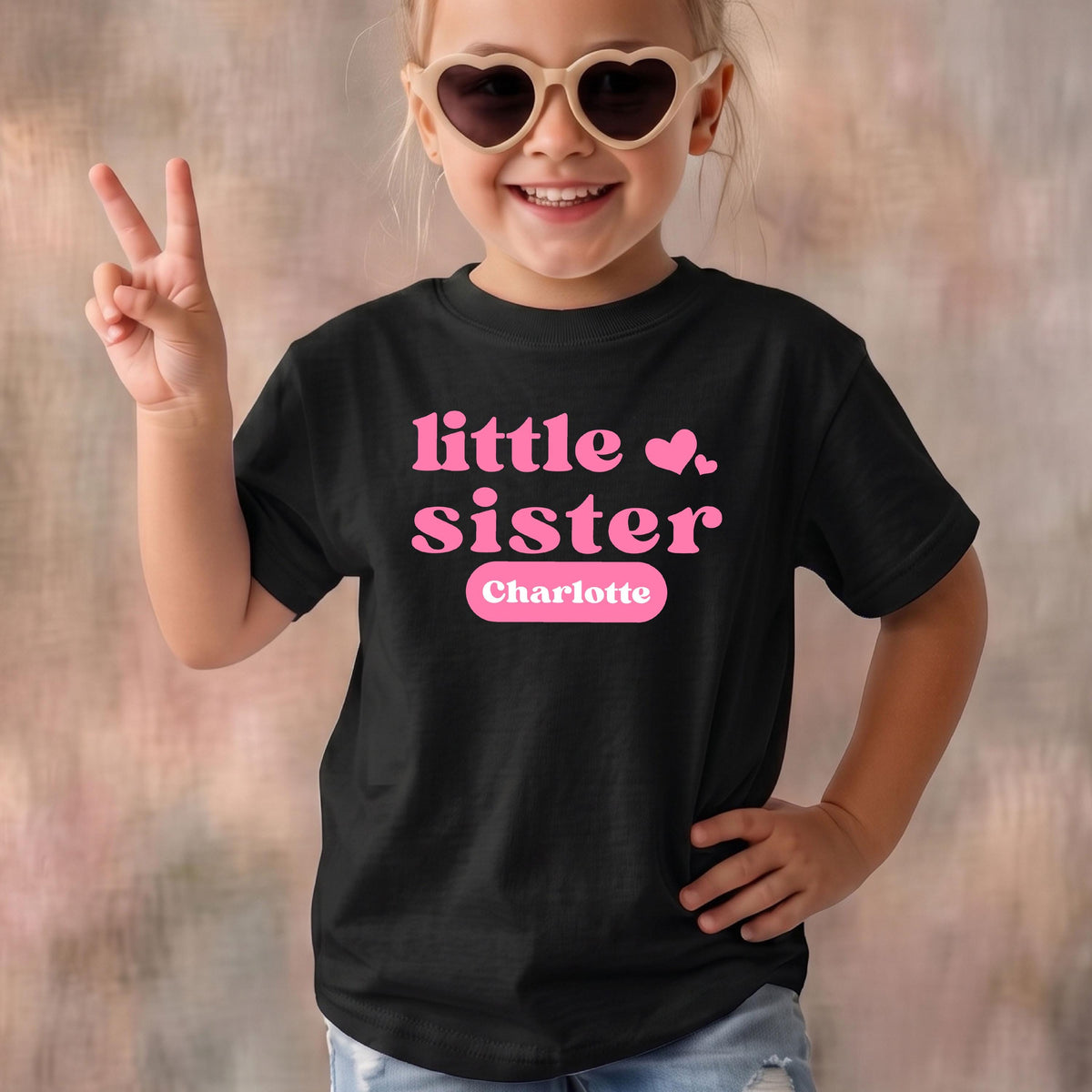 May include: A black short-sleeved t-shirt featuring the pink text "little sister" with two small pink hearts above the word "sister" and the name "Charlotte" printed in white inside a pink rounded rectangular box on the front.