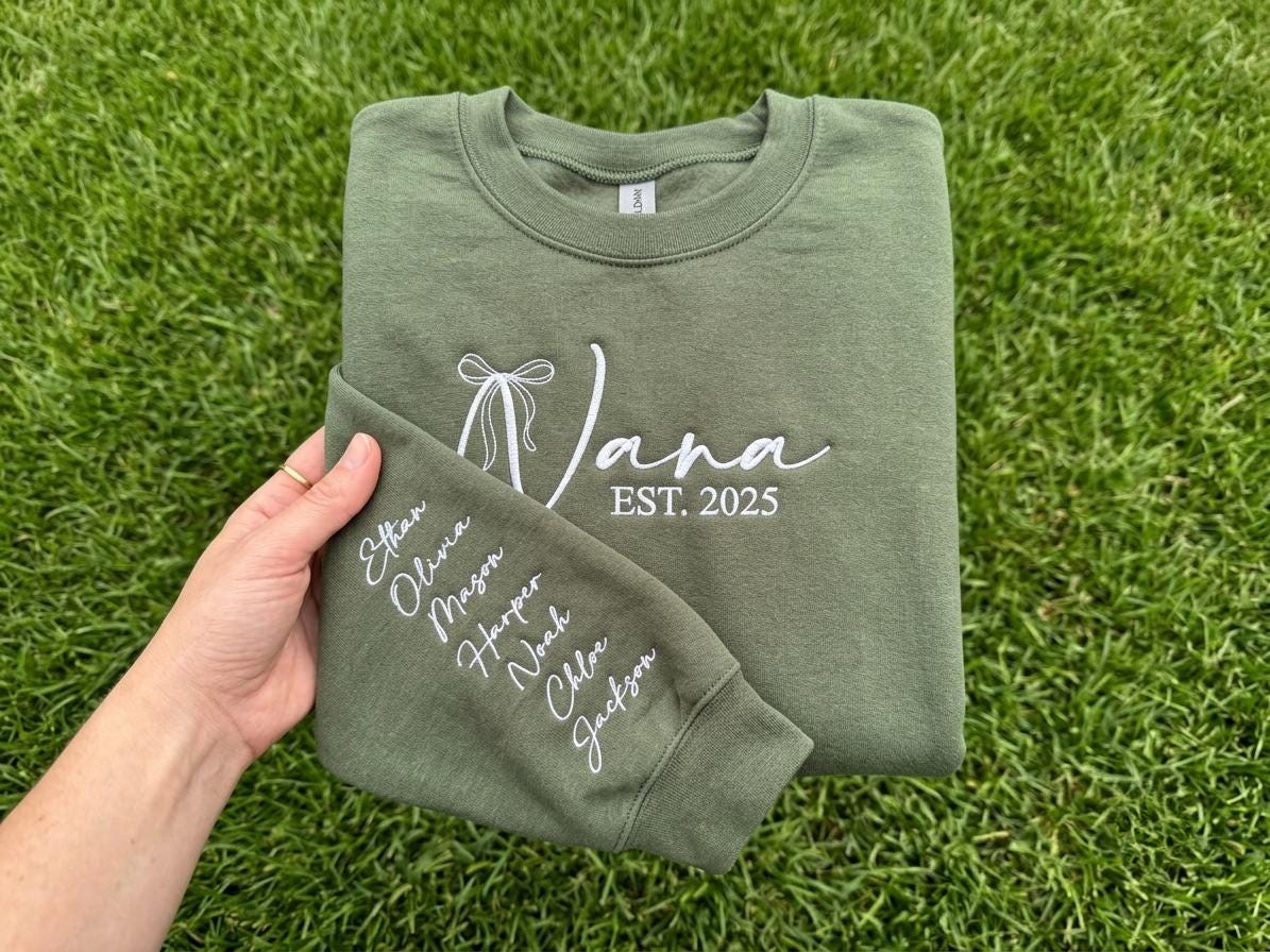 May include: An olive green crewneck sweatshirt featuring white embroidered text on the chest that reads "Nana EST. 2025" with a bow graphic. The sleeve is embroidered with the names Ethan, Olivia, Mason, Harper, Noah, Chloe, and Jackson.