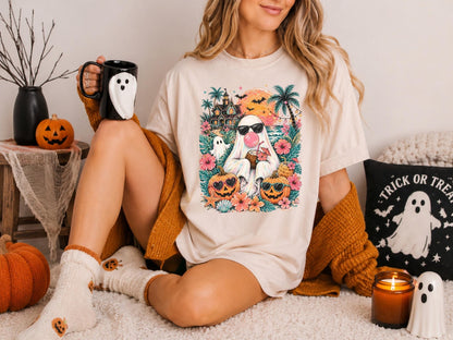 May include: A beige short-sleeve t-shirt featuring a colorful graphic of a ghost wearing sunglasses and blowing a pink bubble, surrounded by tropical palm trees, a haunted house, flowers, and two jack-o'-lanterns with sunglasses.