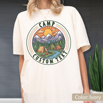 May include: An ivory short-sleeve t-shirt featuring a circular graphic on the chest. The design depicts a mountain landscape with pine trees, two tents, a campfire, and a river under a sun. The text CAMP CUSTOM TEXT is printed in green.