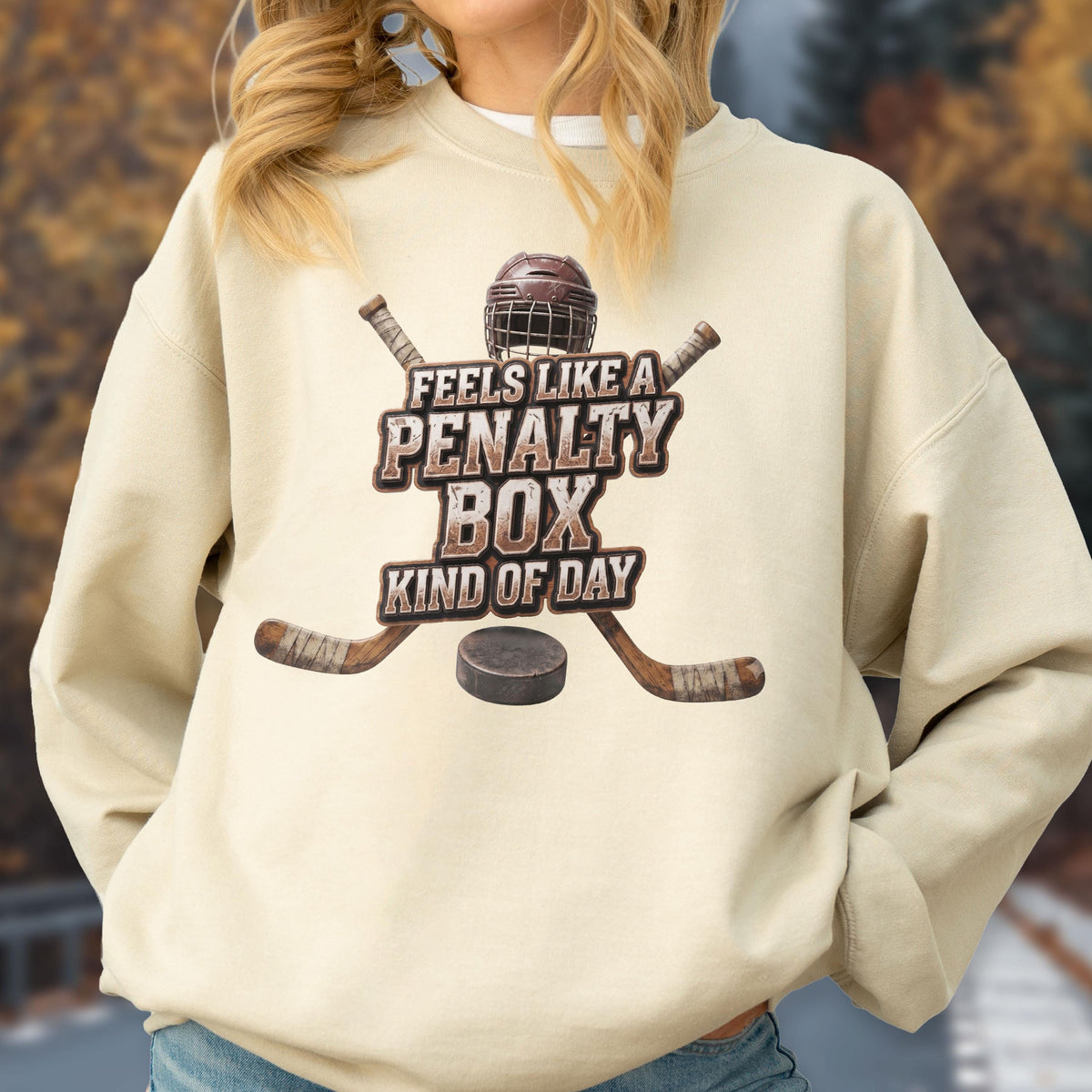 May include: A beige crewneck sweatshirt featuring a graphic of a brown hockey helmet, two crossed hockey sticks, and a black puck. The text reads, "FEELS LIKE A PENALTY BOX KIND OF DAY" in a distressed, brown, block-style font centered on the chest.