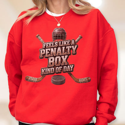 May include: A bright red crewneck sweatshirt featuring a graphic of a hockey helmet, two crossed hockey sticks, and a puck. The text reads, "FEELS LIKE A PENALTY BOX KIND OF DAY," in a distressed brown font centered on the chest of the garment.