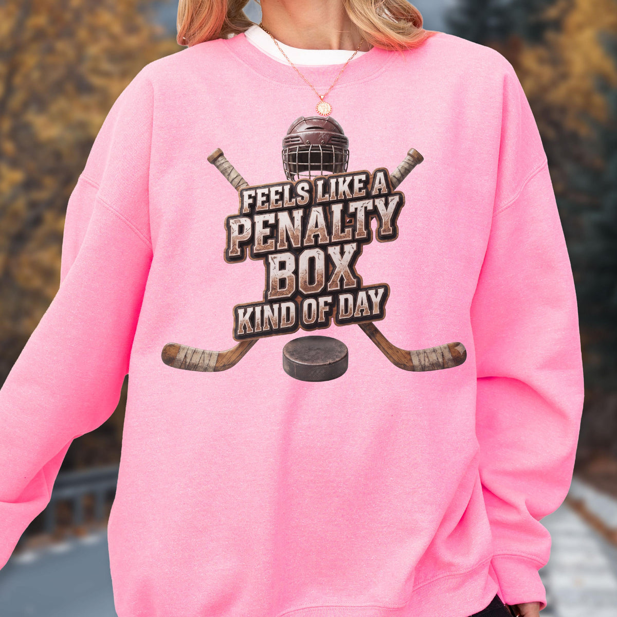 May include: A bright pink crewneck sweatshirt featuring a graphic print of a hockey helmet, two crossed wooden hockey sticks, and a black puck. The text reads FEELS LIKE A PENALTY BOX KIND OF DAY in a distressed brown font centered on the chest.