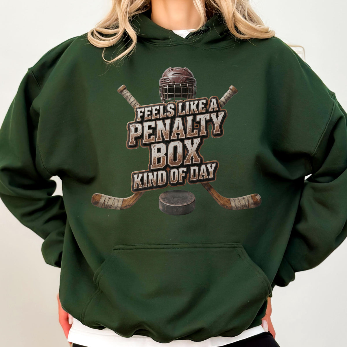 May include: A dark forest green hooded sweatshirt featuring a graphic print of a hockey helmet, two crossed hockey sticks, and a puck. The text reads, "FEELS LIKE A PENALTY BOX KIND OF DAY," centered in a distressed, weathered brown font style.