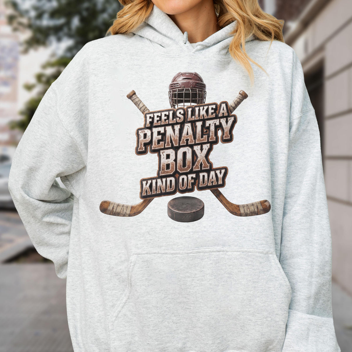May include: A light gray hooded sweatshirt featuring a graphic of a hockey helmet, two crossed hockey sticks, and a puck. The text reads FEELS LIKE A PENALTY BOX KIND OF DAY in a distressed brown font centered on the front of the garment.