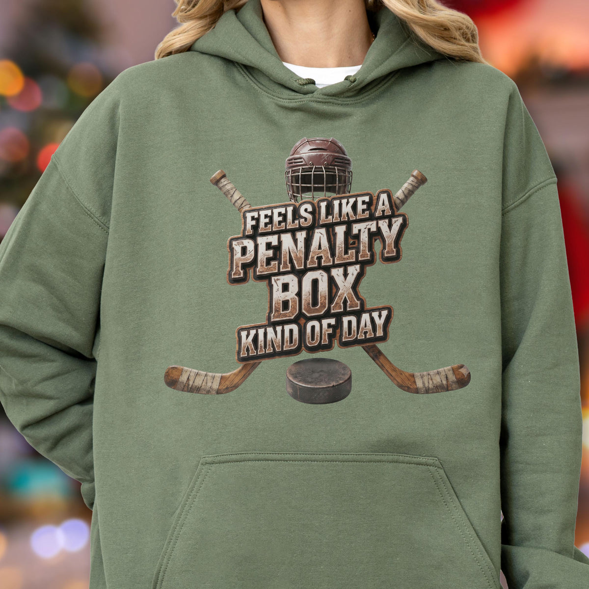 May include: An olive green hooded sweatshirt featuring a graphic print of a hockey helmet, two crossed hockey sticks, and a puck. The text reads FEELS LIKE A PENALTY BOX KIND OF DAY in a distressed brown font centered on the chest of the garment.