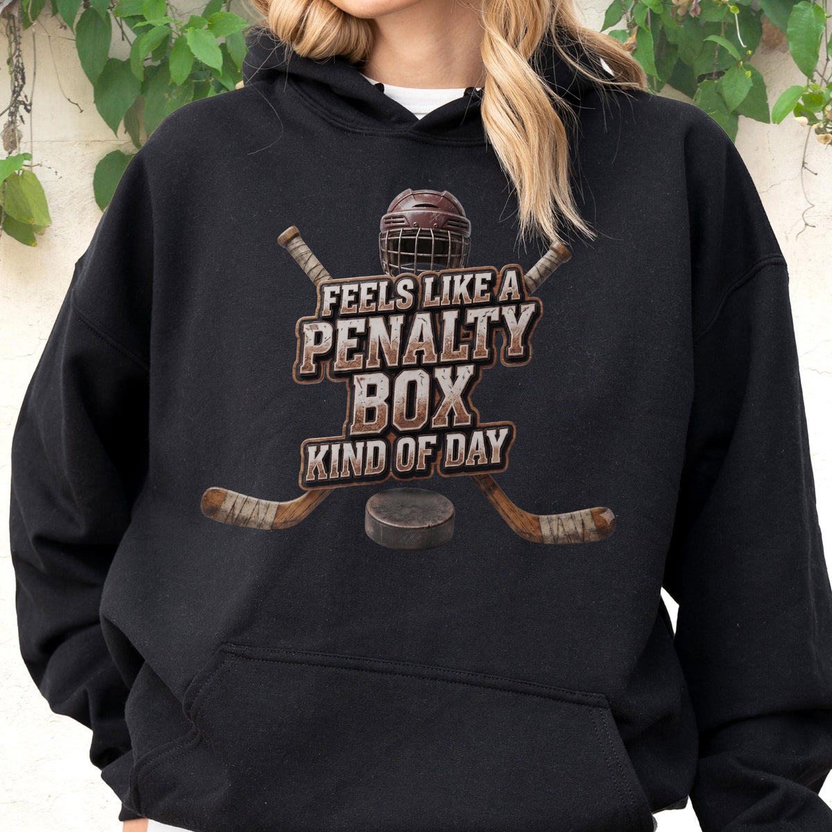 May include: A black hooded sweatshirt featuring a graphic print of a hockey helmet, two crossed hockey sticks, and a puck. The text reads, "FEELS LIKE A PENALTY BOX KIND OF DAY," centered in a distressed, weathered font style on the front chest area.
