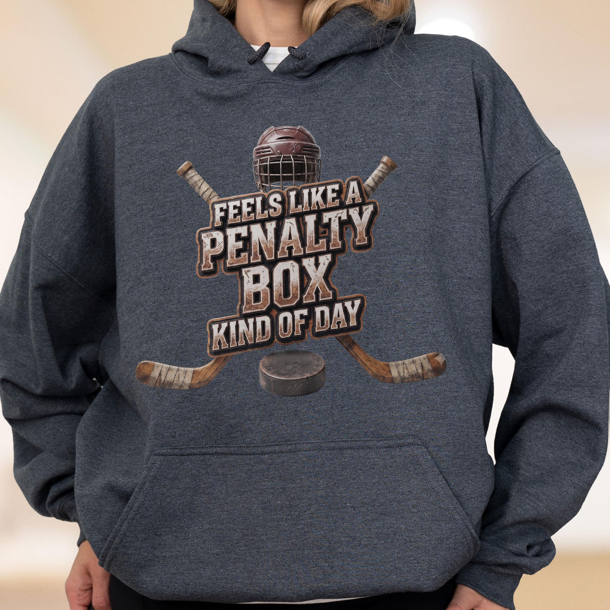 May include: A dark heather gray hooded sweatshirt featuring a graphic print of a hockey helmet, two crossed hockey sticks, and a puck. The text reads FEELS LIKE A PENALTY BOX KIND OF DAY in a distressed, weathered font centered on the chest area.