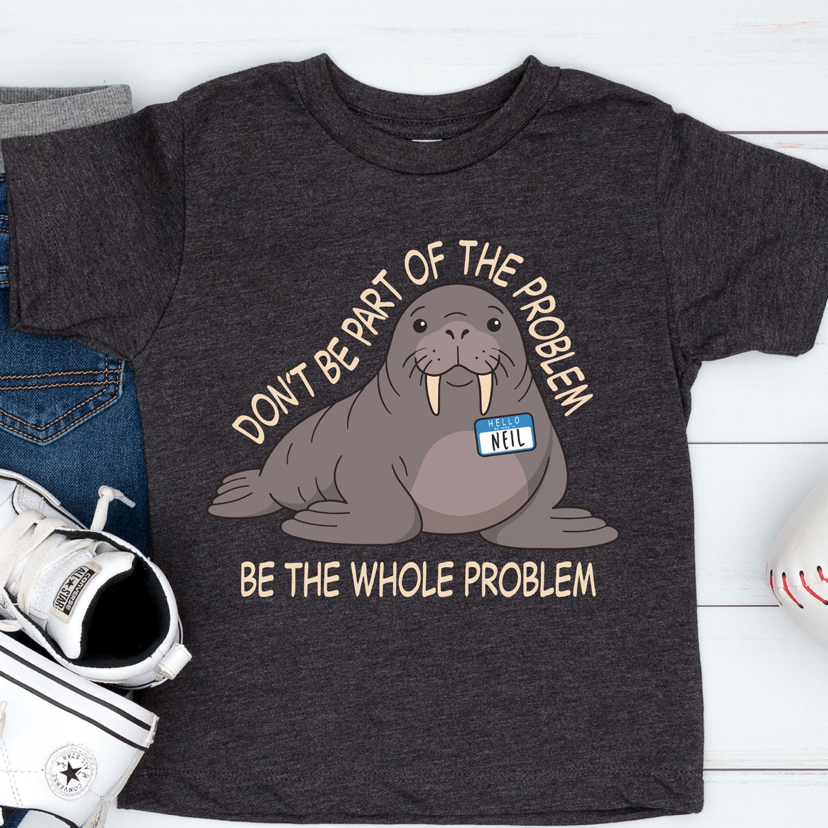 May include: A charcoal gray short-sleeve t-shirt featuring a cartoon walrus with a name tag that reads "HELLO NEIL". The text above the walrus says "DON'T BE PART OF THE PROBLEM" and the text below the walrus says "BE THE WHOLE PROBLEM".