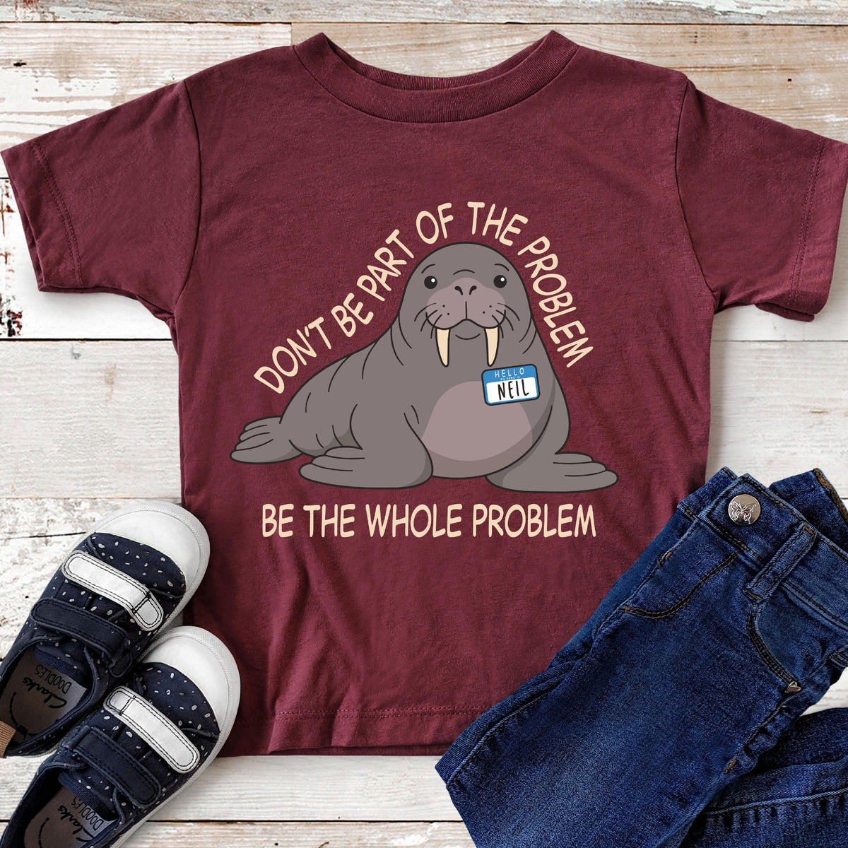 May include: A maroon short-sleeve t-shirt featuring a cartoon walrus with a name tag that reads "HELLO NEIL". The text above and below the walrus reads "DON'T BE PART OF THE PROBLEM" and "BE THE WHOLE PROBLEM" in a light cream-colored font.