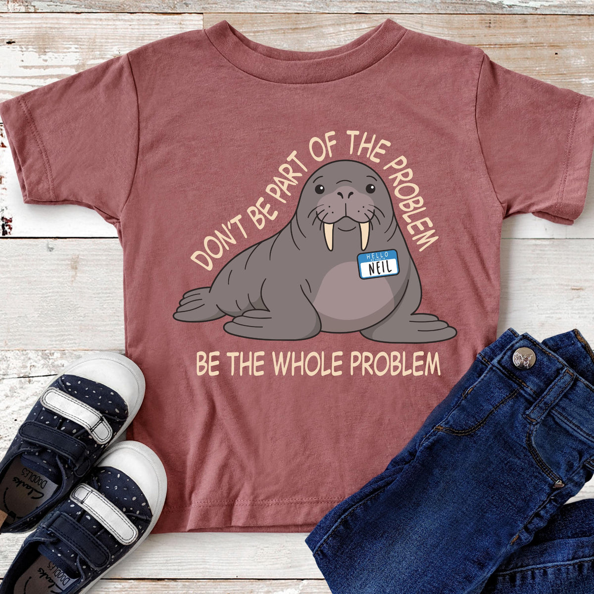 May include: A mauve short-sleeved t-shirt featuring a graphic of a grey walrus wearing a blue name tag that reads "HELLO NEIL". The text "DON'T BE PART OF THE PROBLEM" is arched above the walrus, and "BE THE WHOLE PROBLEM" is printed below.