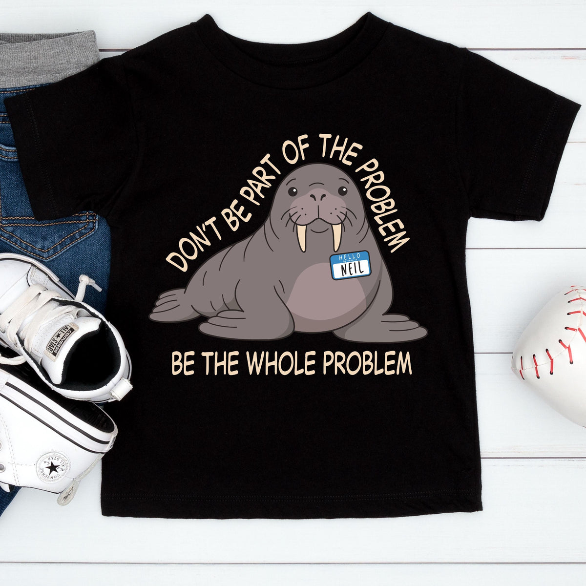 May include: A black t-shirt featuring a cartoon walrus with a name tag that reads "HELLO NEIL". The text above the walrus says "DON'T BE PART OF THE PROBLEM" and the text below the walrus says "BE THE WHOLE PROBLEM" in a light beige font.