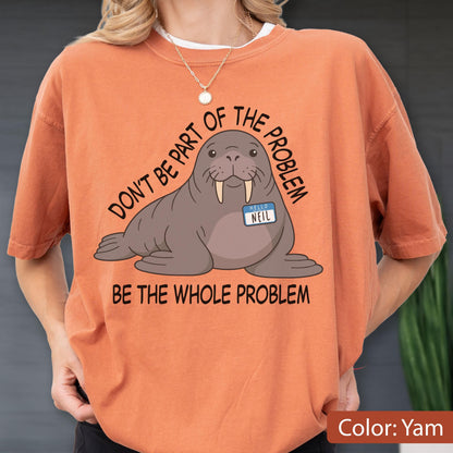 May include: A burnt orange crew neck t-shirt featuring a cartoon walrus illustration with a name tag that reads "HELLO NEIL". The text above the walrus says "DON'T BE PART OF THE PROBLEM" and the text below reads "BE THE WHOLE PROBLEM".