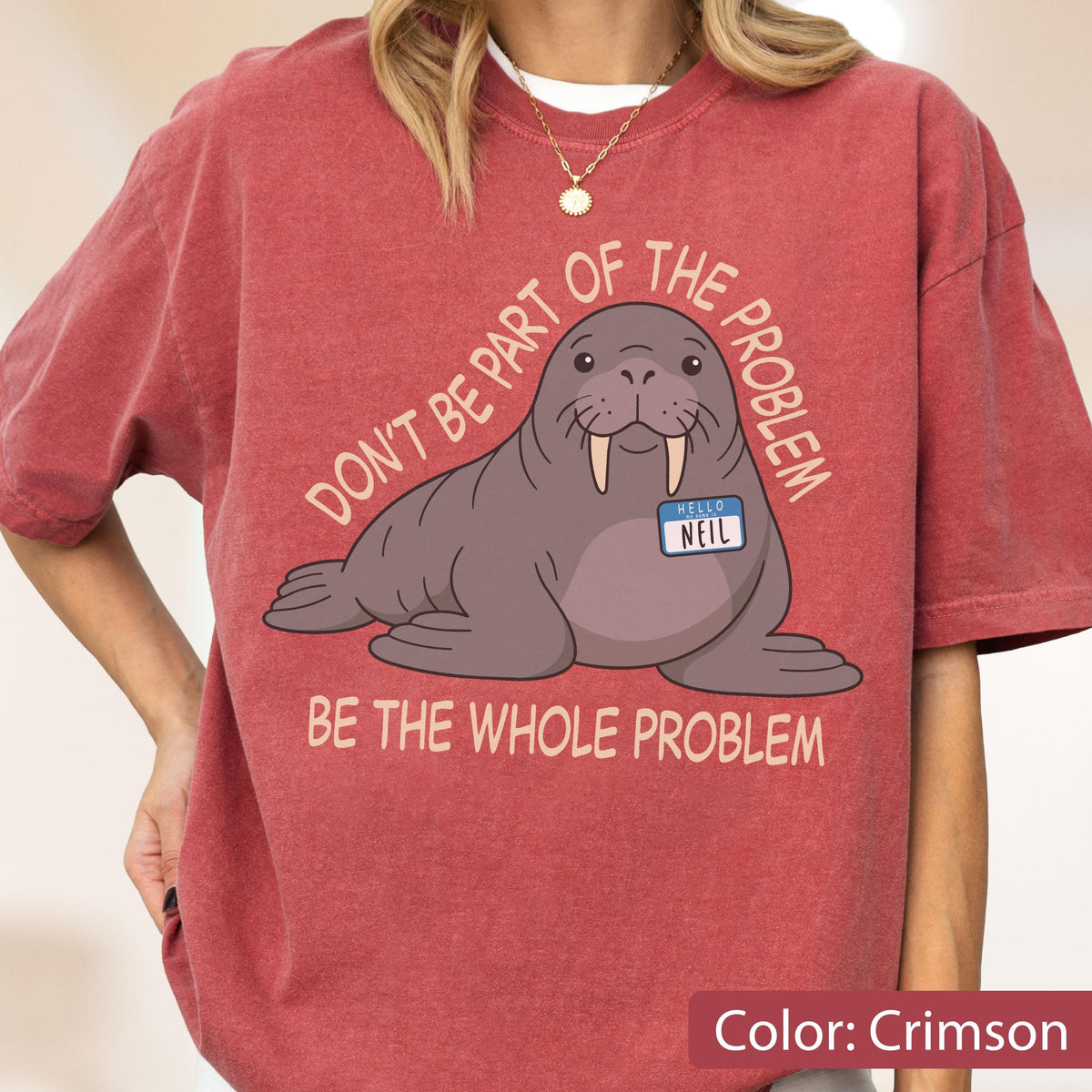 May include: A crimson short-sleeve t-shirt featuring a graphic of a brown walrus wearing a name tag that reads "HELLO NEIL". The text "DON'T BE PART OF THE PROBLEM" is arched above the walrus, and "BE THE WHOLE PROBLEM" is printed below it.