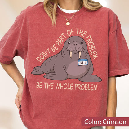May include: A crimson short-sleeve t-shirt featuring a graphic of a brown walrus wearing a name tag that reads "HELLO NEIL". The text "DON'T BE PART OF THE PROBLEM" is arched above the walrus, and "BE THE WHOLE PROBLEM" is printed below it.