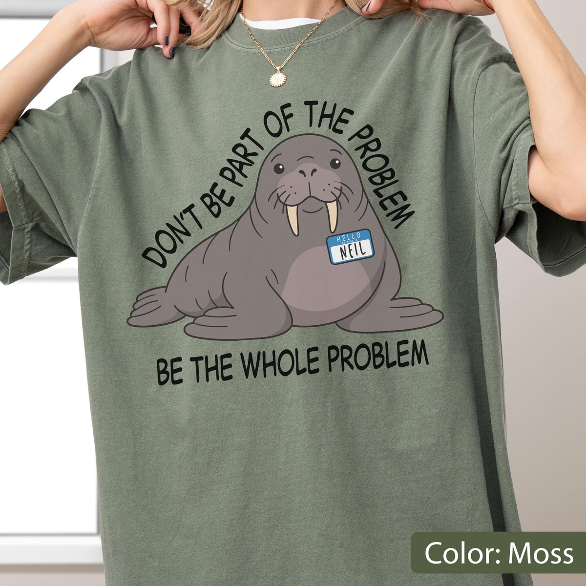 May include: A moss green t-shirt featuring a graphic of a grey walrus with tusks wearing a blue name tag that reads HELLO NEIL. The text surrounding the walrus reads DON'T BE PART OF THE PROBLEM and BE THE WHOLE PROBLEM in black lettering.