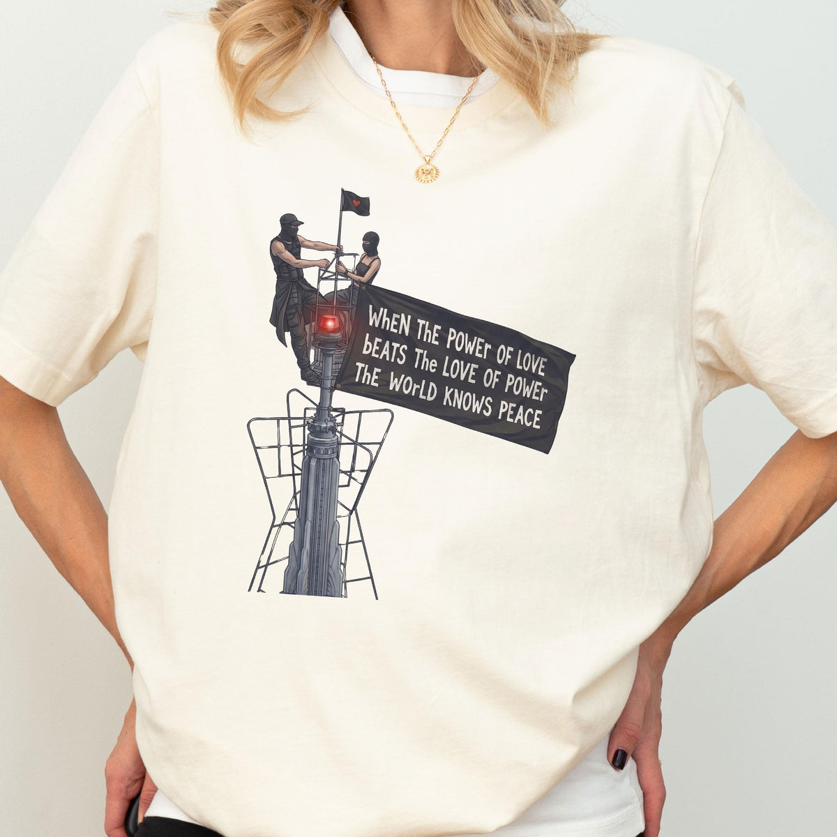 May include: A cream-colored t-shirt featuring a graphic of two figures atop a structure holding a black flag. The flag displays the text: WHEN THE POWER OF LOVE BEATS THE LOVE OF POWER THE WORLD KNOWS PEACE. The design includes a red light detail.