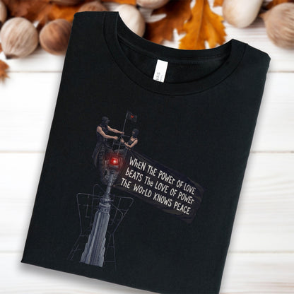 May include: A black t-shirt featuring a graphic of two figures standing atop a tall structure, one holding a flag with the text: WHEN THE POWER OF LOVE BEATS THE LOVE OF POWER THE WORLD KNOWS PEACE. The design includes a red light and tower details.