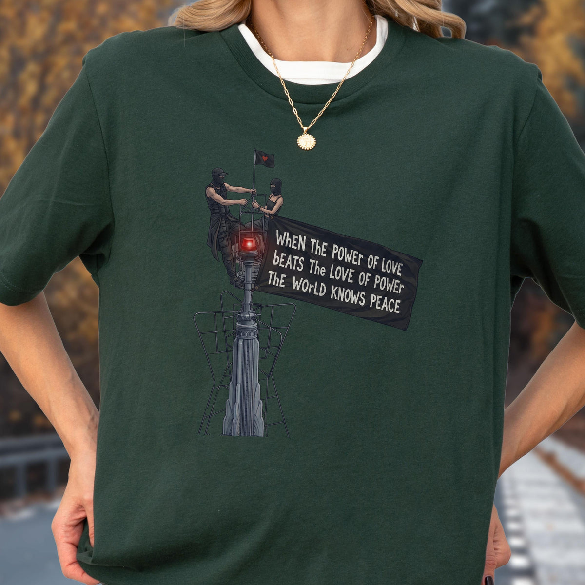 May include: A dark forest green short-sleeve t-shirt featuring a graphic of two figures atop a tall structure holding a black flag. The flag displays the text: WHEN THE POWER OF LOVE BEATS THE LOVE OF POWER THE WORLD KNOWS PEACE in white lettering.
