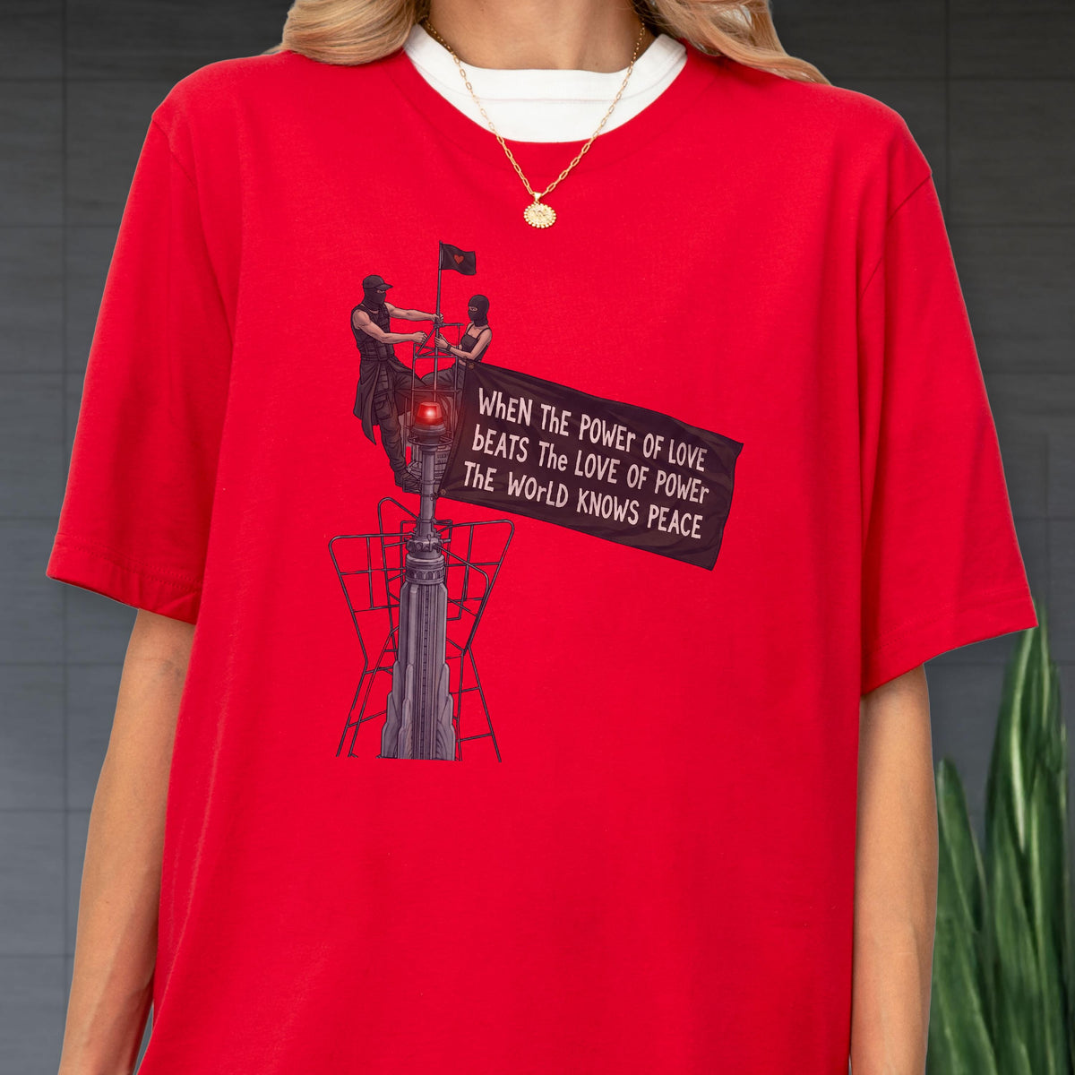 May include: A bright red short-sleeved t-shirt featuring a graphic of two figures atop a tall structure holding a black banner. The text on the banner reads: WHEN THE POWER OF LOVE BEATS THE LOVE OF POWER THE WORLD KNOWS PEACE. A gold chain necklace is worn.