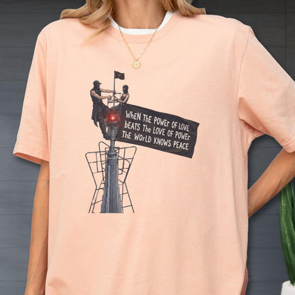 May include: A peach-colored t-shirt featuring a graphic of two figures atop a tall structure holding a black flag. The flag displays the text: When The POWER of LOVE bEATS The LOVE of POWEr The WoRLD KNOWS PEACE. The design includes a red light.