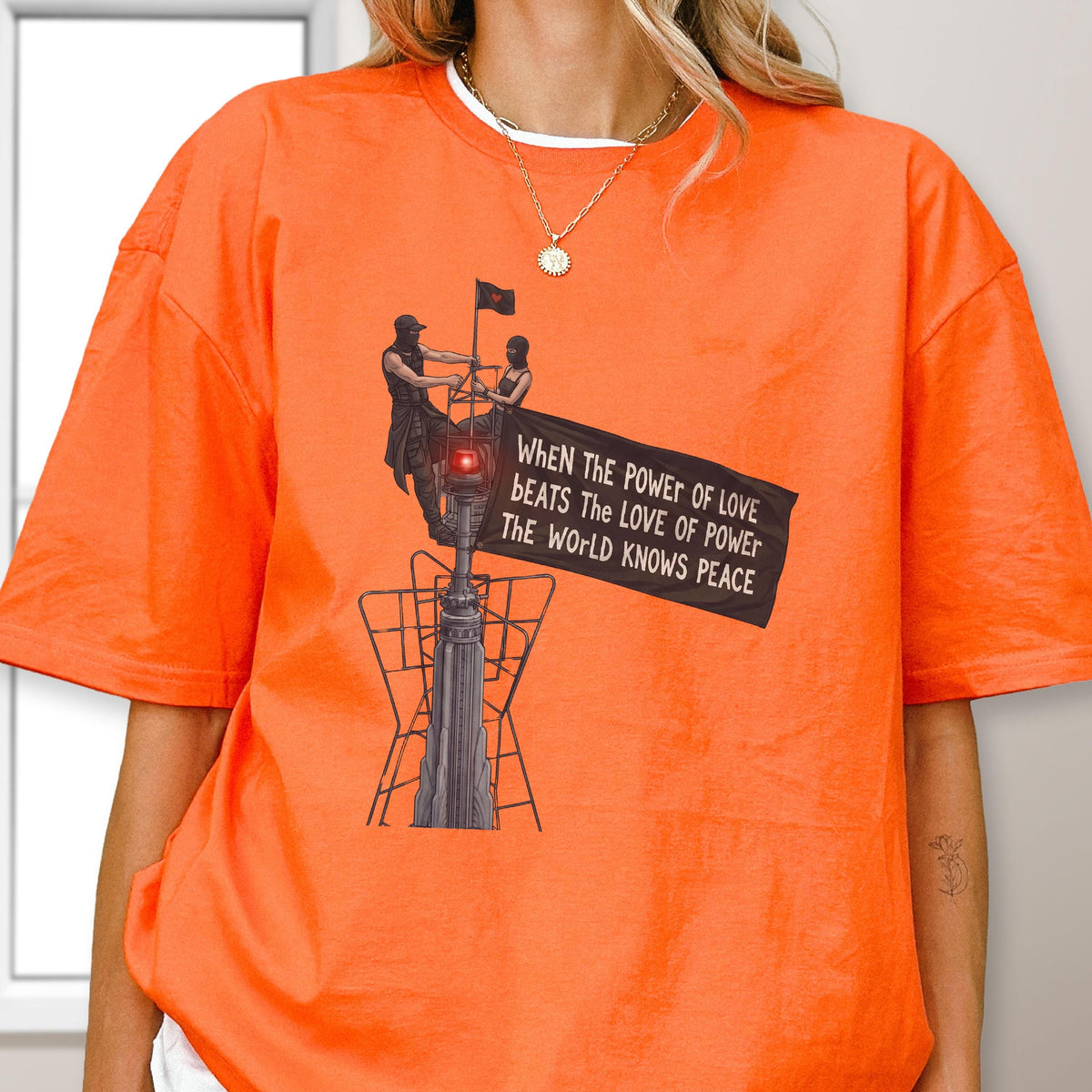 May include: An orange short-sleeved t-shirt featuring a graphic of two figures atop a tall structure holding a black flag. The flag displays the text: WHEN THE POWER OF LOVE BEATS THE LOVE OF POWER THE WORLD KNOWS PEACE. The design includes a red light.