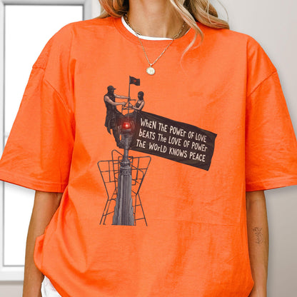 May include: An orange short-sleeved t-shirt featuring a graphic of two figures atop a tall structure holding a black flag. The flag displays the text: WHEN THE POWER OF LOVE BEATS THE LOVE OF POWER THE WORLD KNOWS PEACE. The design includes a red light.
