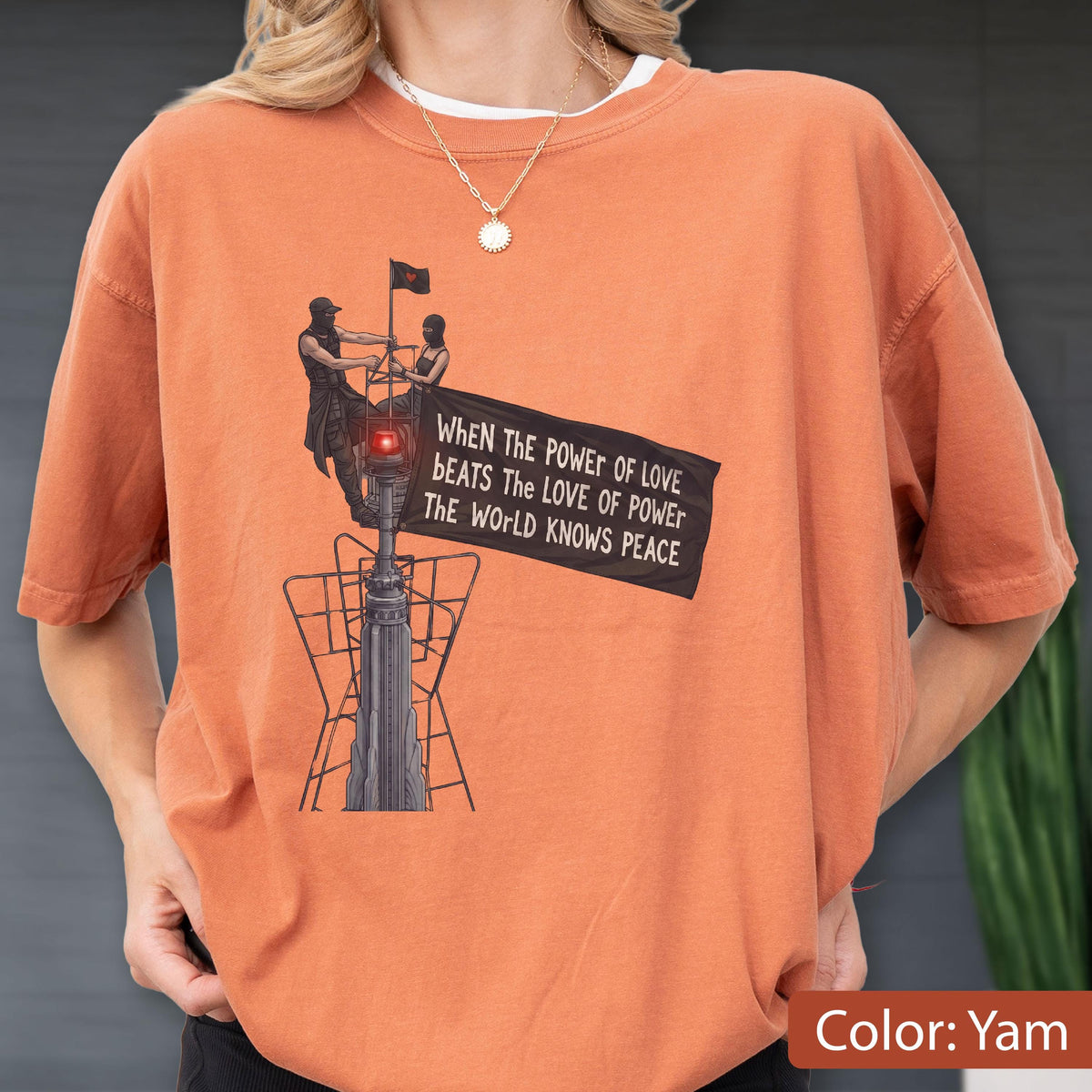 May include: A yam-colored t-shirt featuring a graphic of two figures atop a tall structure holding a black flag. The flag displays the text: When the power of love beats the love of power the world knows peace. The design includes a red light.