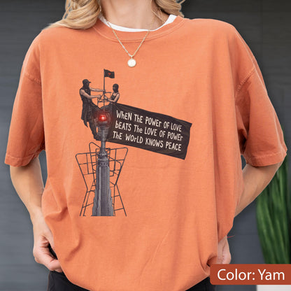 May include: A yam-colored t-shirt featuring a graphic of two figures atop a tall structure holding a black flag. The flag displays the text: When the power of love beats the love of power the world knows peace. The design includes a red light.