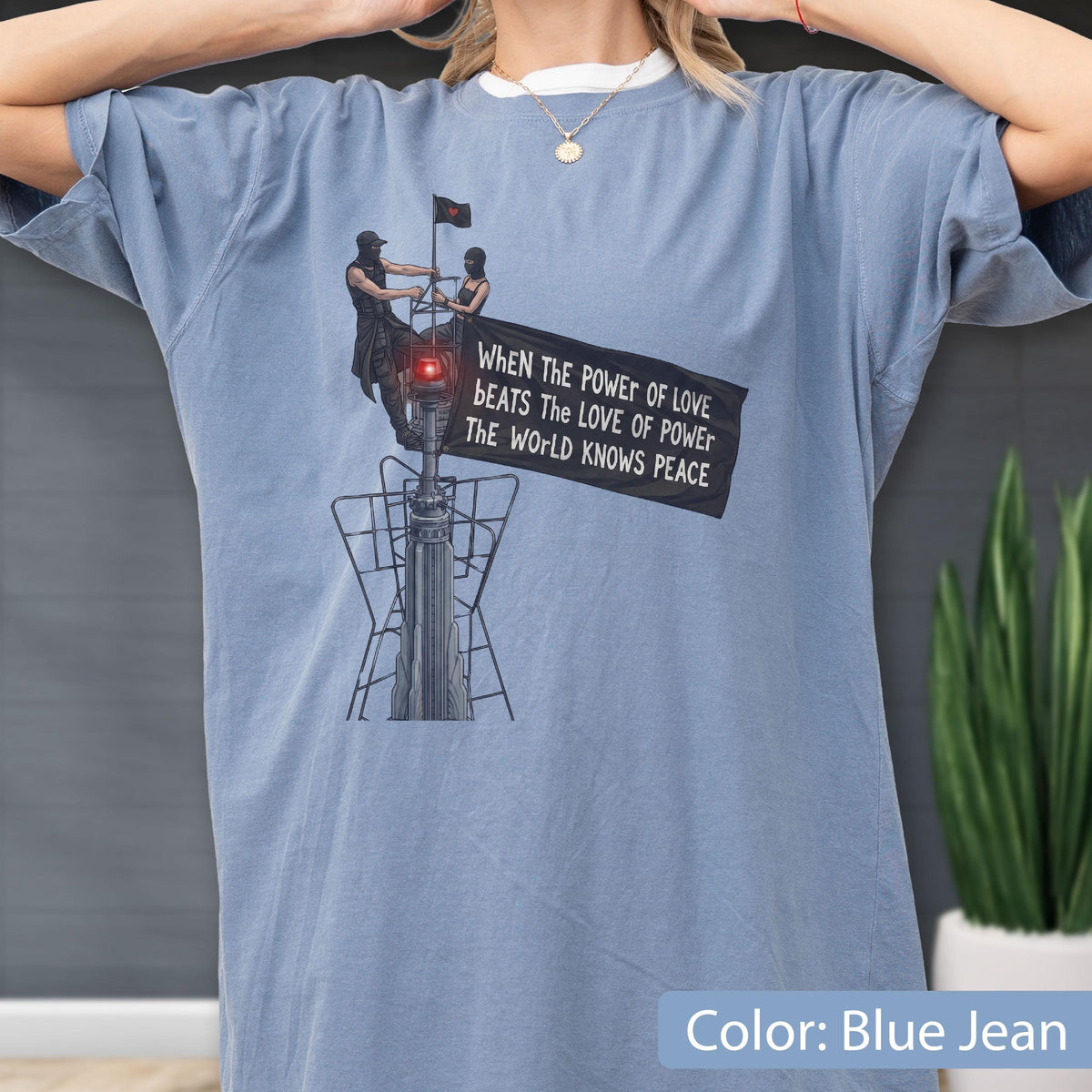 May include: A blue jean colored t-shirt featuring a graphic of two figures atop a tower holding a black flag. The flag displays the text: WHEN THE POWER OF LOVE BEATS THE LOVE OF POWER THE WORLD KNOWS PEACE. The design includes a red light detail.