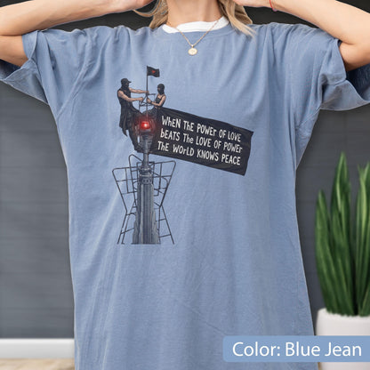May include: A blue jean colored t-shirt featuring a graphic of two figures atop a tower holding a black flag. The flag displays the text: WHEN THE POWER OF LOVE BEATS THE LOVE OF POWER THE WORLD KNOWS PEACE. The design includes a red light detail.