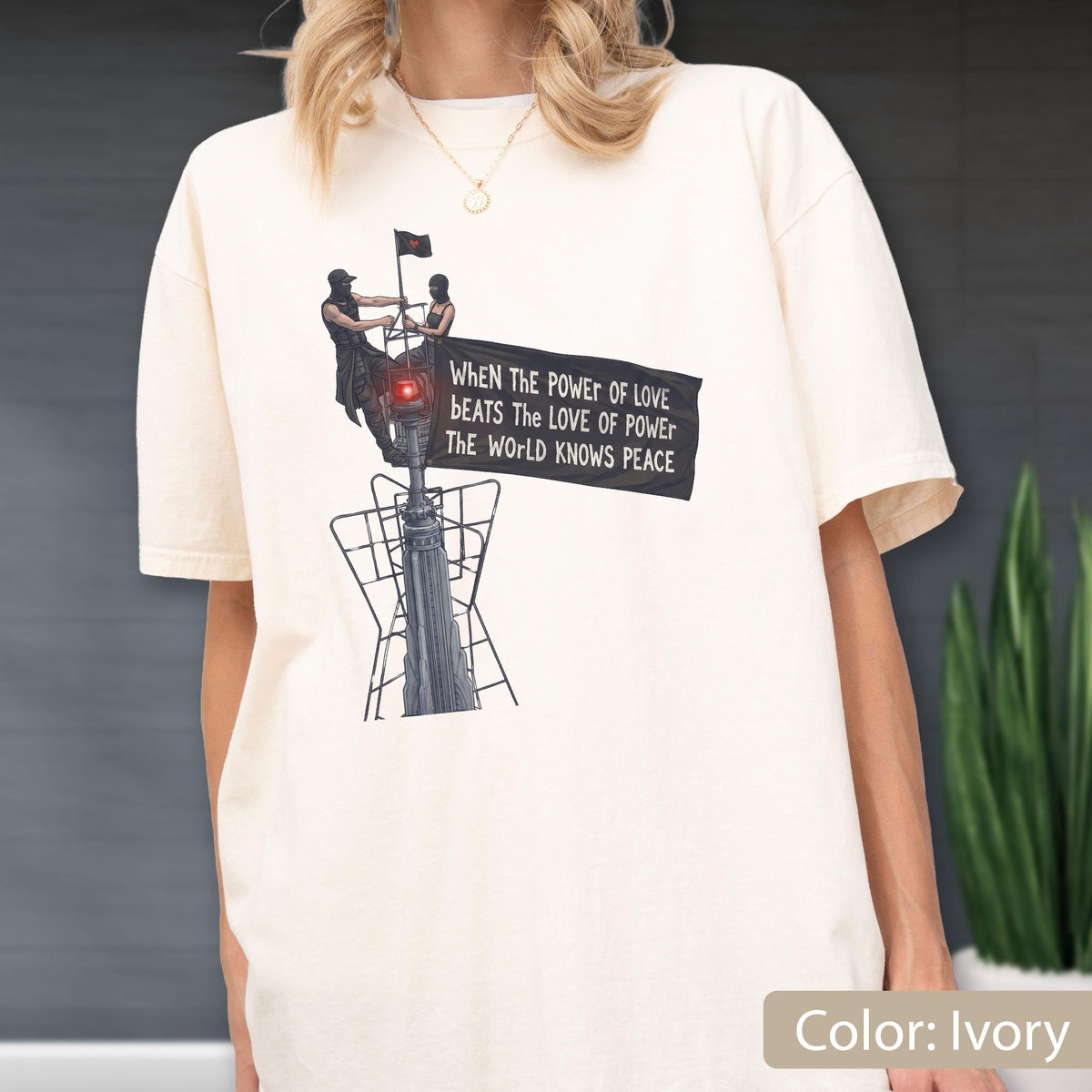 May include: An ivory short-sleeve t-shirt featuring a graphic of two figures atop a tall structure holding a black banner. The banner displays the text: When the power of love beats the love of power the world knows peace. The design includes a red light.
