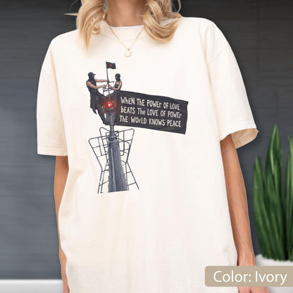 May include: An ivory short-sleeve t-shirt featuring a graphic of two figures atop a tall structure holding a black banner. The banner displays the text: When the power of love beats the love of power the world knows peace. The design includes a red light.
