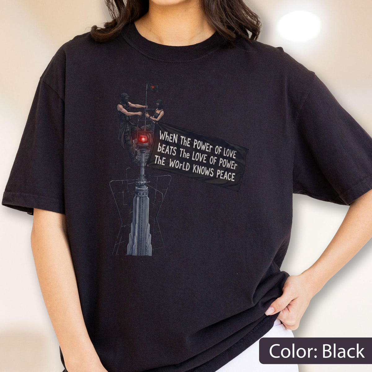 May include: A black short-sleeved t-shirt featuring a graphic of two figures atop a tall structure holding a banner that reads: When the power of love beats the love of power the world knows peace. The design includes a glowing red light element.