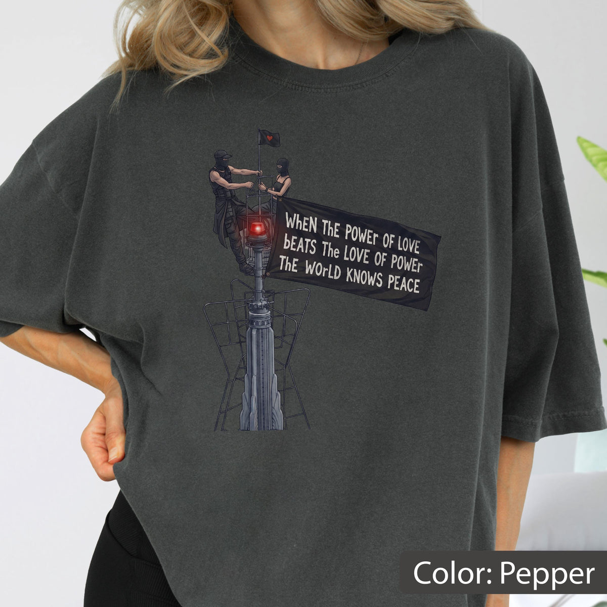 May include: A pepper-colored t-shirt featuring a graphic of two figures atop a tower holding a black flag. The flag displays the text: WHEN THE POWER OF LOVE BEATS THE LOVE OF POWER THE WORLD KNOWS PEACE. The design includes a small red light.