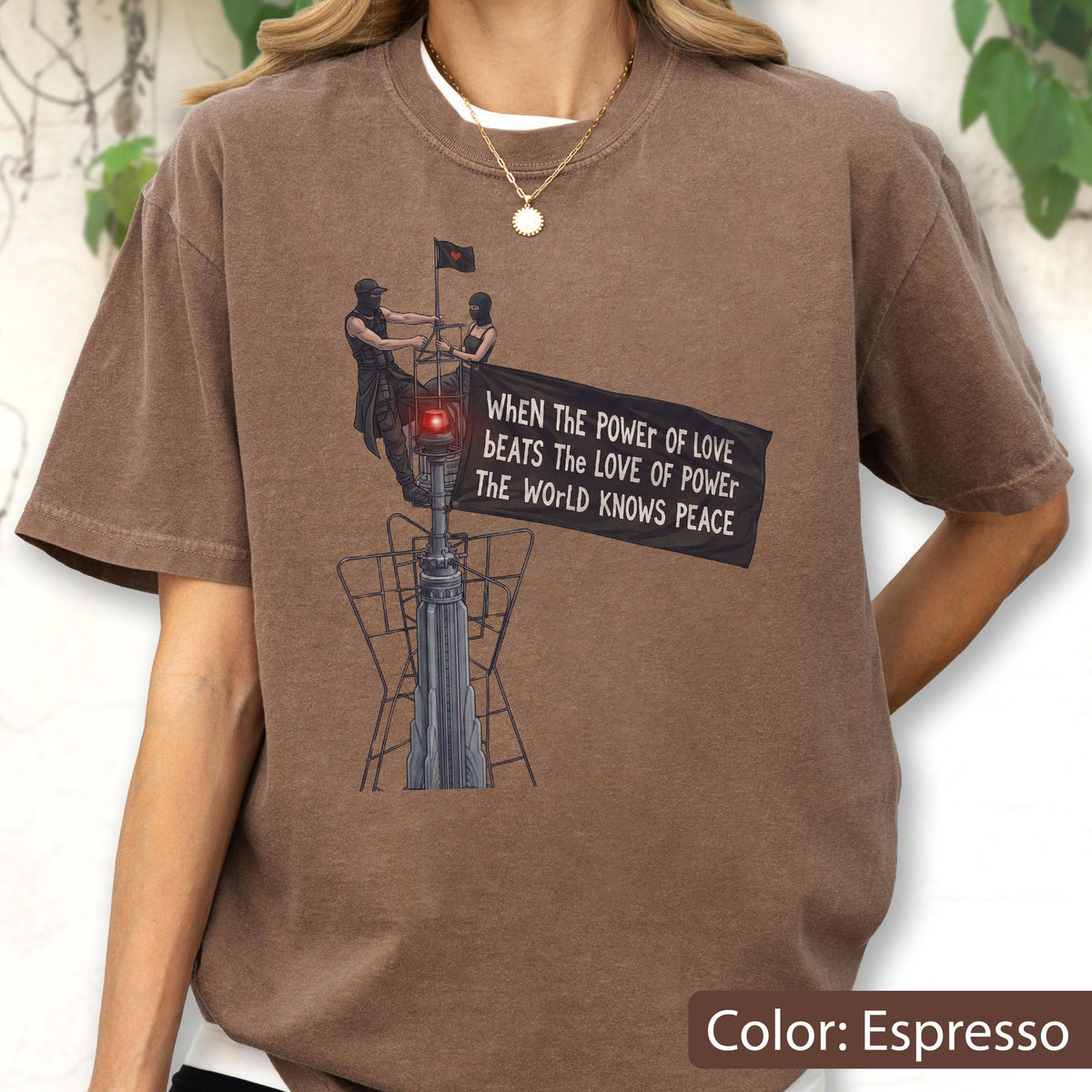 May include: An espresso-colored t-shirt featuring a graphic of two figures atop a tower structure holding a black flag. The flag displays the text: When The Power Of Love Beats The Love Of Power The World Knows Peace. The design includes a red light.