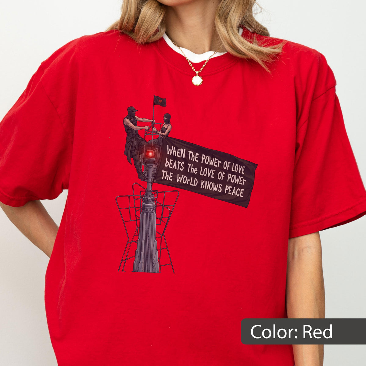 May include: A bright red short-sleeved t-shirt featuring a graphic print of two figures atop a tall structure holding a banner that reads: When The Power Of Love Beats The Love Of Power The World Knows Peace. The design includes a small red light.