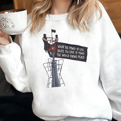 May include: A white crewneck sweatshirt featuring a graphic print of two figures atop a tall structure holding a black flag. The text on the flag reads: WHEN THE POWER OF LOVE BEATS THE LOVE OF POWER THE WORLD KNOWS PEACE. The design includes a red light.