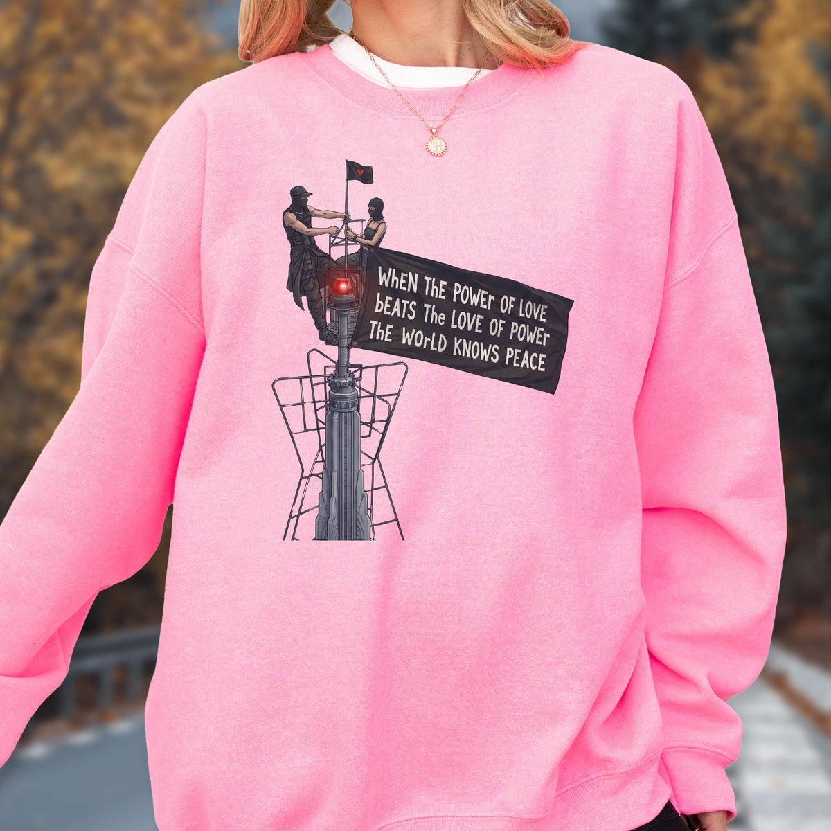 May include: A bright pink crewneck sweatshirt featuring a graphic print of two figures atop a tall structure holding a black banner. The text on the banner reads: When the power of love beats the love of power the world knows peace.