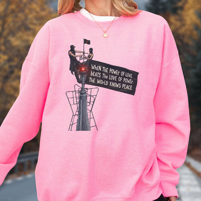 May include: A bright pink crewneck sweatshirt featuring a graphic print of two figures atop a tall structure holding a black banner. The text on the banner reads: When the power of love beats the love of power the world knows peace.