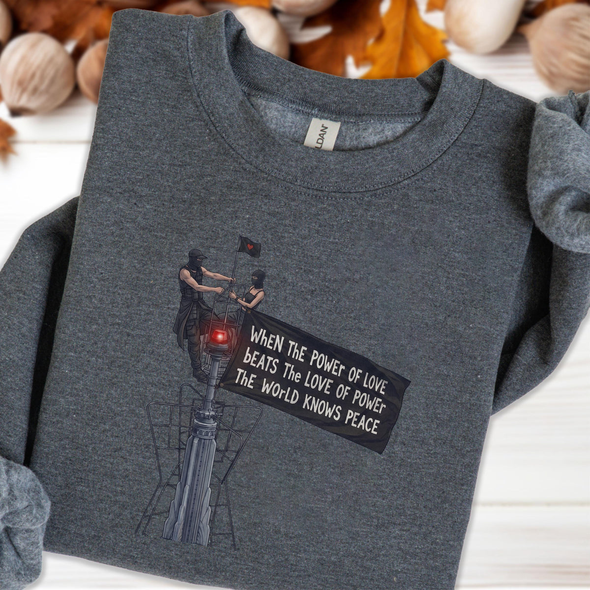 May include: A dark heather gray crewneck sweatshirt featuring a graphic print of two figures standing on a tall structure, holding a black flag with the text: When the power of love beats the love of power the world knows peace.
