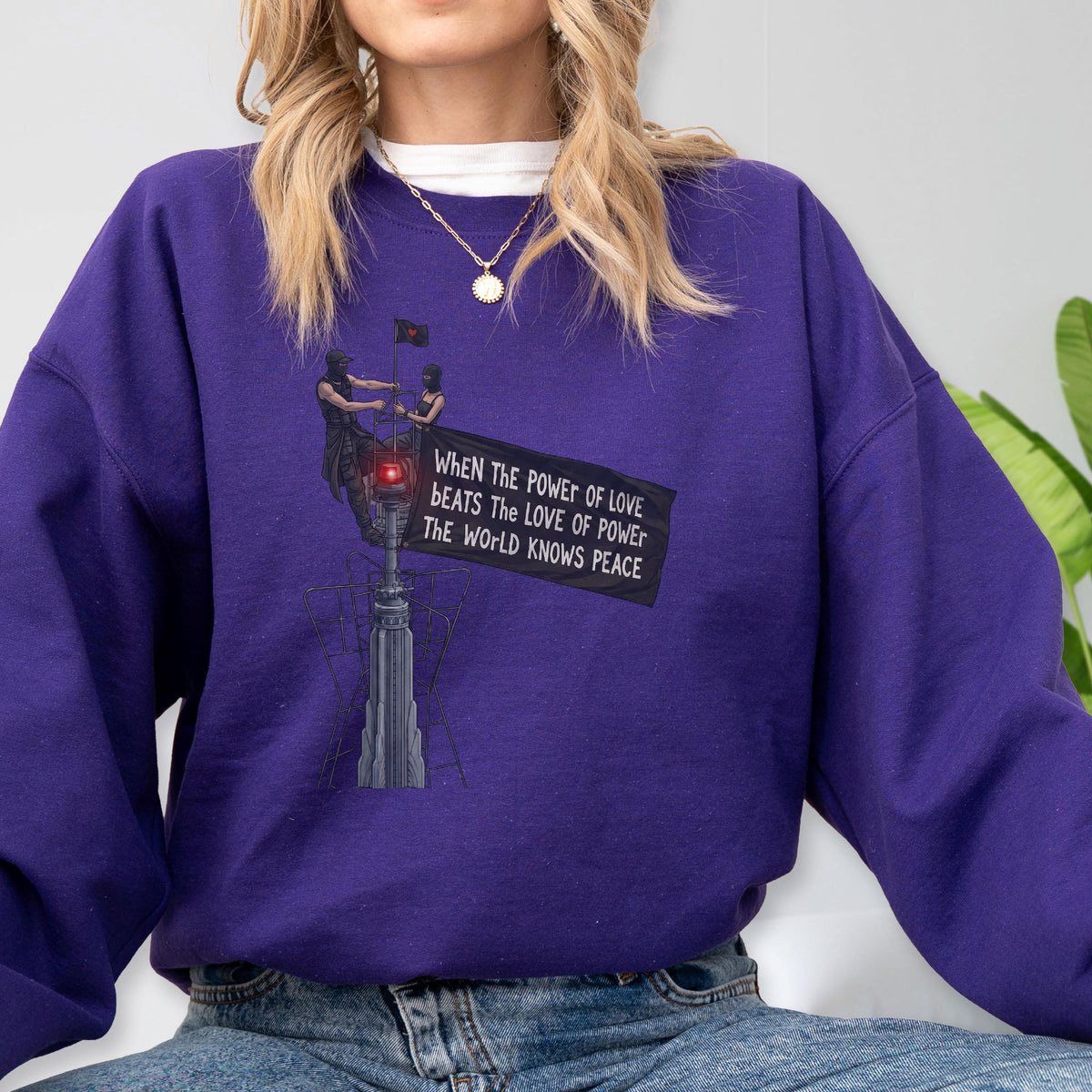 May include: A purple long-sleeve sweatshirt featuring a graphic print of two figures atop a tall structure holding a banner that reads: When the power of love beats the love of power the world knows peace. The design includes a red light detail.