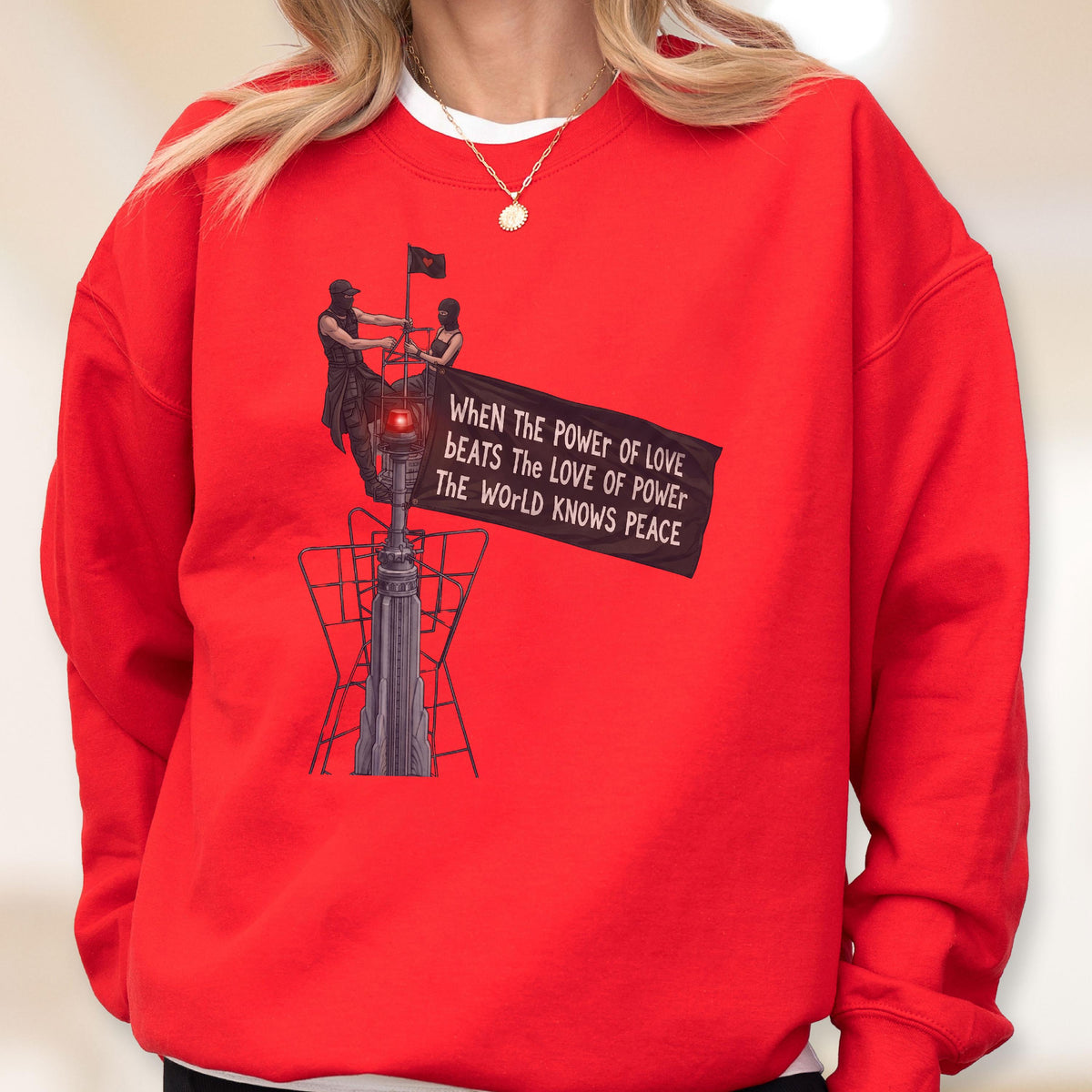May include: A bright red crewneck sweatshirt featuring a graphic illustration of two figures standing atop a tall structure, holding a black flag with the text: When The Power Of Love Beats The Love Of Power The World Knows Peace.