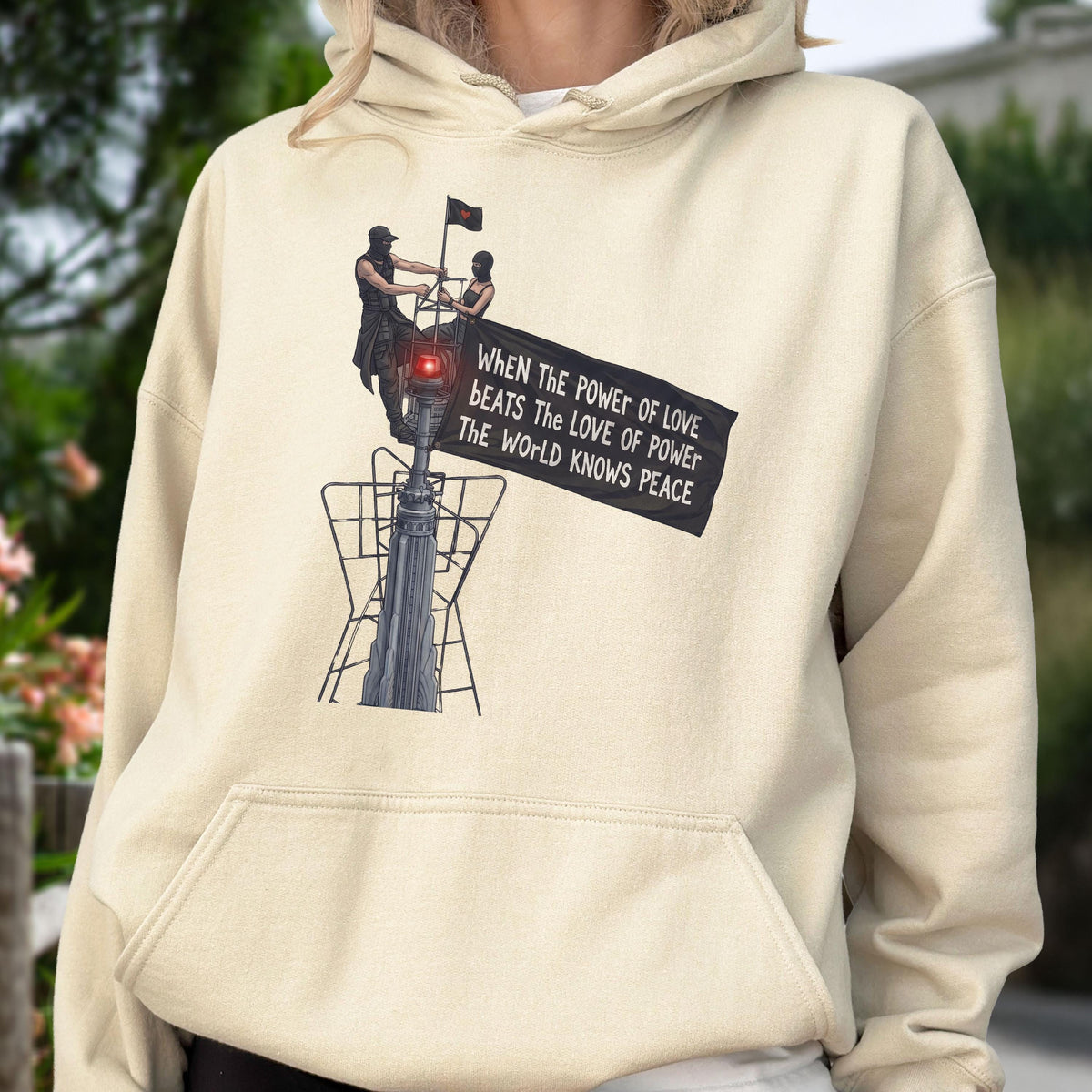 May include: A cream-colored hoodie featuring a graphic print of two figures standing atop a tall structure, holding a black banner that reads: When the power of love beats the love of power the world knows peace. The hoodie has a front kangaroo pocket.