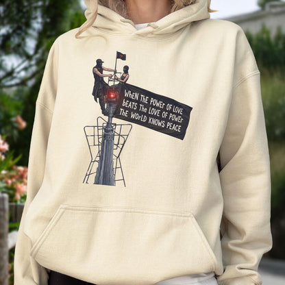 May include: A cream-colored hoodie featuring a graphic print of two figures standing atop a tall structure, holding a black banner that reads: When the power of love beats the love of power the world knows peace. The hoodie has a front kangaroo pocket.