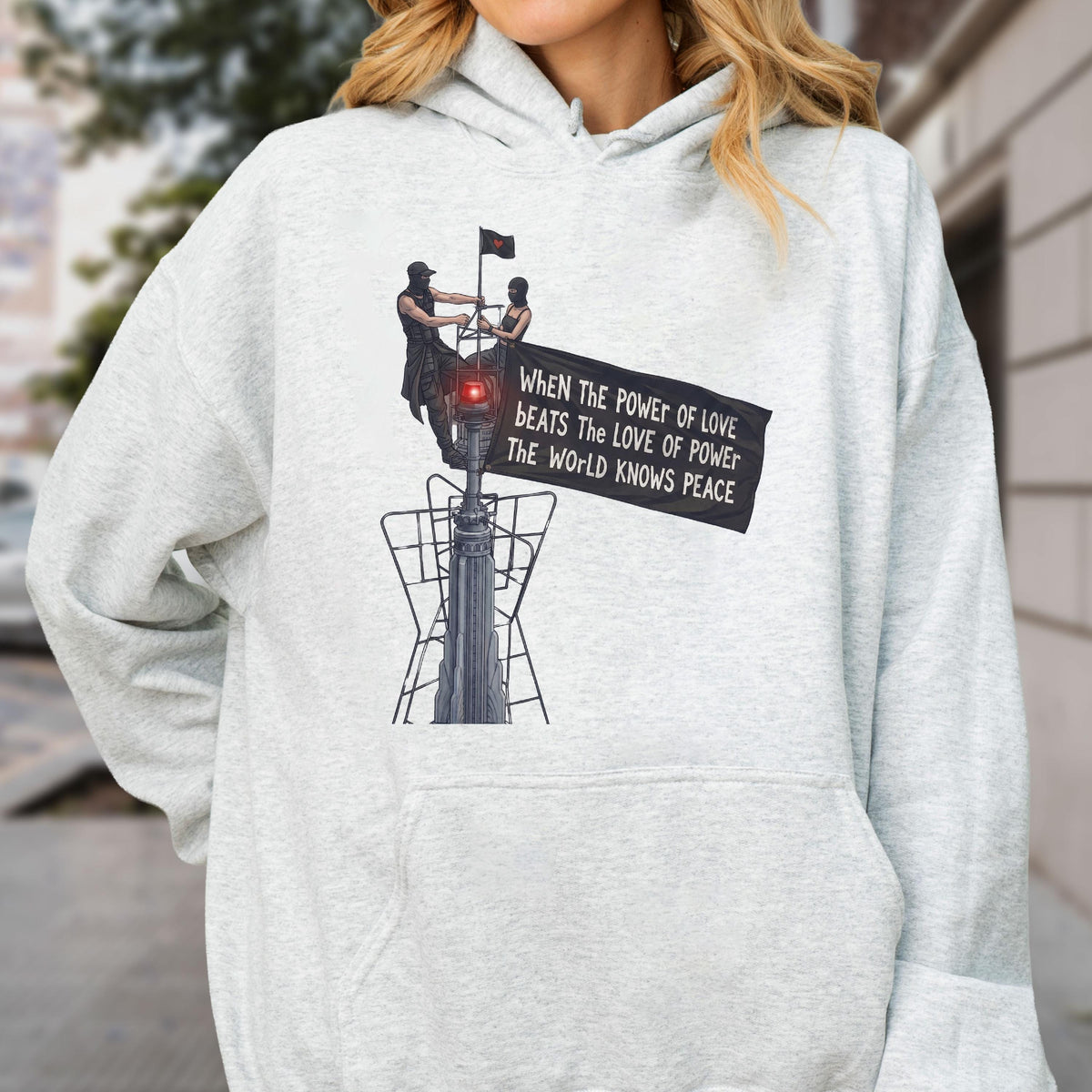 May include: A light gray heathered hoodie featuring a graphic print of two figures atop a tall structure holding a black banner. The banner displays the text: When the power of love beats the love of power the world knows peace.