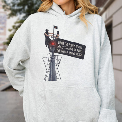 May include: A light gray heathered hoodie featuring a graphic print of two figures atop a tall structure holding a black banner. The banner displays the text: When the power of love beats the love of power the world knows peace.