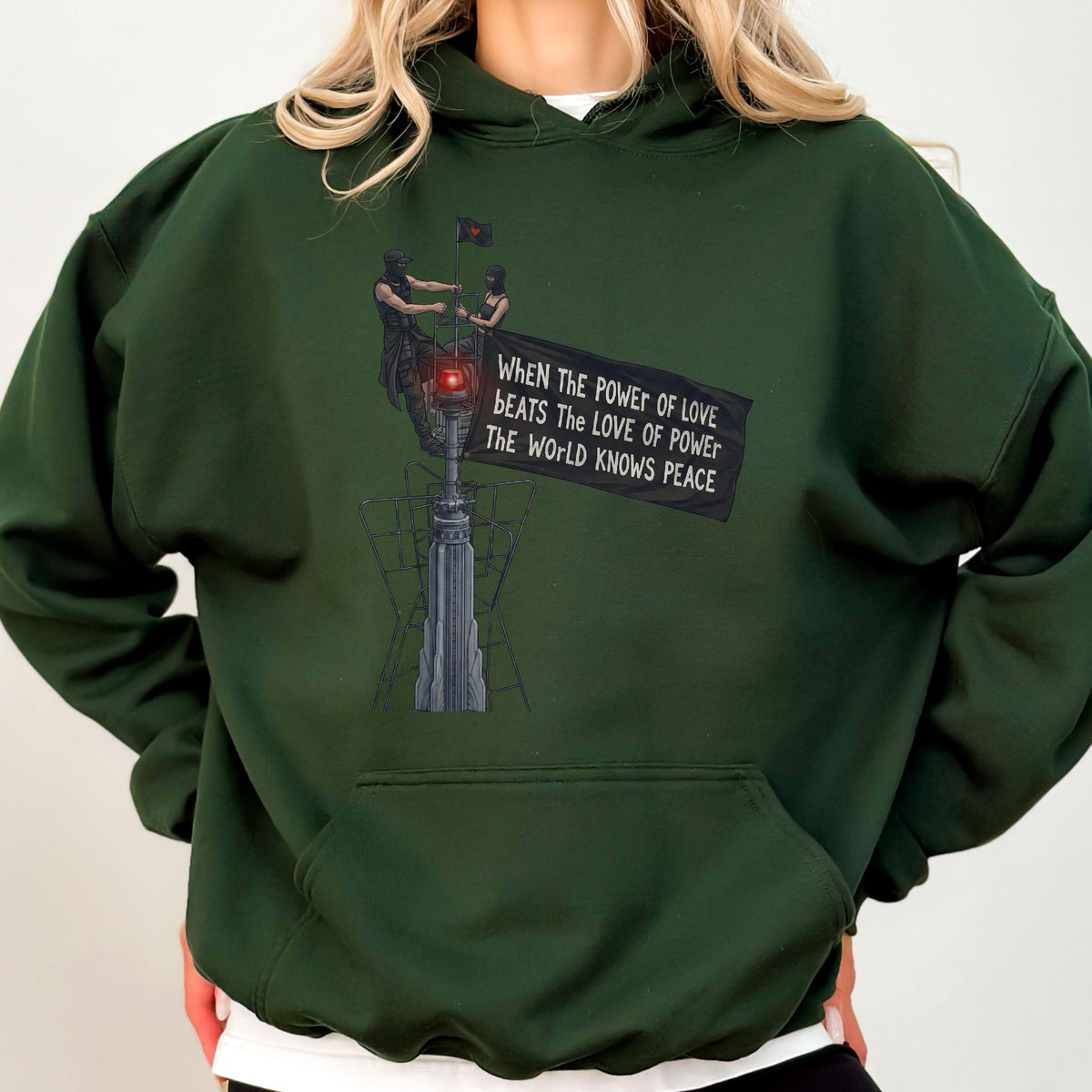 May include: A dark forest green hooded sweatshirt featuring a graphic print of two figures atop a tall structure holding a black flag. The flag displays the text: When The Power Of Love Beats The Love Of Power The World Knows Peace.