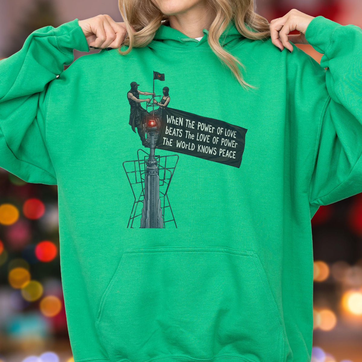 May include: A bright green hooded sweatshirt featuring a graphic print of two figures atop a tall structure holding a banner that reads, "WHEN THE POWER OF LOVE BEATS THE LOVE OF POWER THE WORLD KNOWS PEACE." The hoodie has long sleeves and a front pocket.